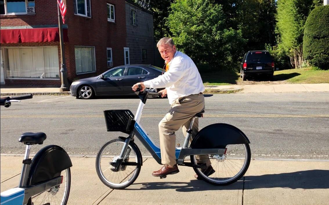A man rides an electric-assist bicycle down a street lined with buildings