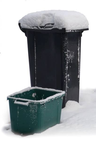 A snow-topped black trash can stands next to a snowy green recycling bin 